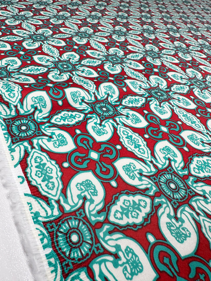 A close-up of Super Cheap Fabrics’ Cotton Sateen - Mural - 150cm fabric, showcasing intricate teal and white floral geometric designs on a red background, perfect for stylish décor with its bold, symmetrical repeating motif.
