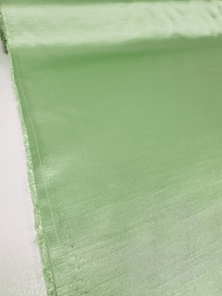 A close-up of the Shantung Satin - Foam Green - 150cm by Super Cheap Fabrics, displaying its silky texture with a slightly reflective surface. The edges of this lightweight satin fabric are unfinished and slightly frayed, showcasing the material's sheen and texture. The backdrop is a plain, light-colored surface.