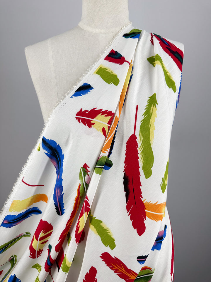 A white mannequin displays Super Cheap Fabrics' Bamboo Rayon - Feather White (150cm), a beginner-friendly sewing fabric with a colorful feather pattern in red, green, blue, and yellow on a plain grey background.