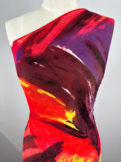 A dress form showcasing the Super Cheap Fabrics' Printed Mesh - Infawave - 153cm, which features a vibrant, abstract-patterned polyester fabric in hues of red, yellow, purple, and black. The one-shoulder design and the colorful brushstroke-like pattern create a dynamic and artistic look that accentuates the lightweight material.