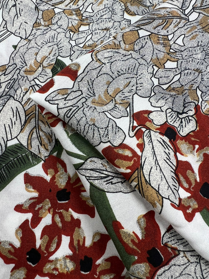 A close-up of the Super Cheap Fabrics' Printed Rayon - XL Flora - 145cm, featuring intricately detailed flowers in stunning shades of red, white, and gold with lush green leaves. The fabric beautifully showcases a blend of blooming floral and foliage patterns, creating a rich, textured design.