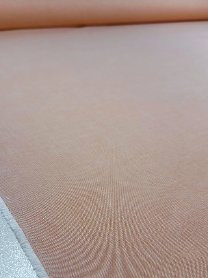 A close-up of Oxford Cotton - Sunburst - 146cm by Super Cheap Fabrics shows a smooth, light peach fabric unrolled on a flat surface, with the roll’s edge visible in the background.