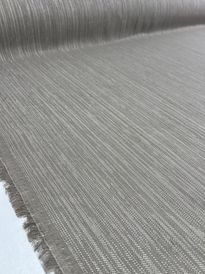 Close-up of a roll of gray Upholstery Twill - Natural - 147cm from Super Cheap Fabrics with subtle, linear texture. The durable fabric appears smooth and tightly woven, with frayed edges visible along the bottom and left sides. The image captures the fine details and texture, ideal for furniture upholstery needs.