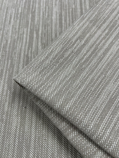 Close-up of folded gray Upholstery Twill - Natural - 147cm by Super Cheap Fabrics with a subtle linear pattern. The fabric showcases a texture comprising thin, light-colored lines running diagonally, creating a modern, sleek aesthetic perfect for durable furniture upholstery.