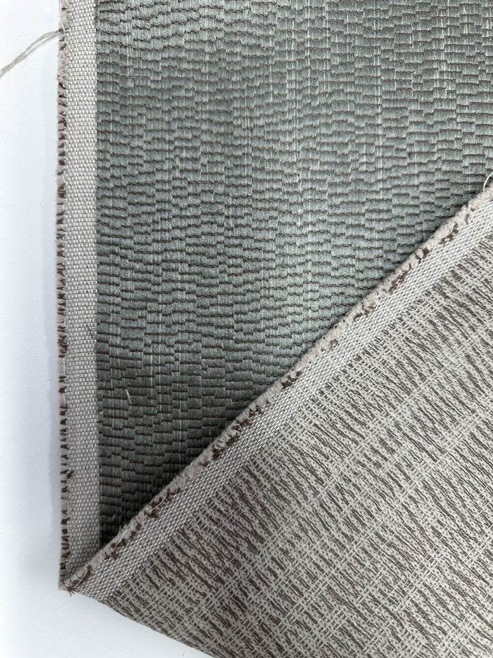 A close-up of Super Cheap Fabrics' Cut Pile Jacquard Upholstery - Oscar Gunmetal (142cm) shows its textured gray-green surface, with a folded corner revealing the woven backing and stitched edge on a white background.
