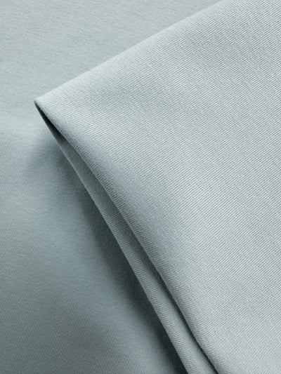 A close-up of Super Cheap Fabrics' Cotton Lycra - Winter Sky (164cm), featuring light gray fabric with a smooth, lightly textured surface. The stretchy material is neatly folded, showing sharp creases and overlapping layers.