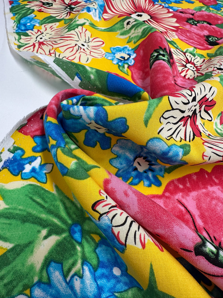 A close-up of Super Cheap Fabrics’ Designer Rayon - Sunset Bloom (145cm), showing a vibrant floral print with red, pink, blue, and white flowers on yellow, accented by green leaves. The fabric is slightly folded and draped.