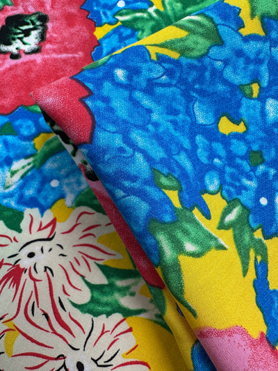 Close-up of Super Cheap Fabrics’ Designer Rayon - Sunset Bloom (145cm), featuring a bold floral pattern with blue, pink, and white flowers plus green leaves on a yellow background. The vibrant fabric appears slightly folded in the image.