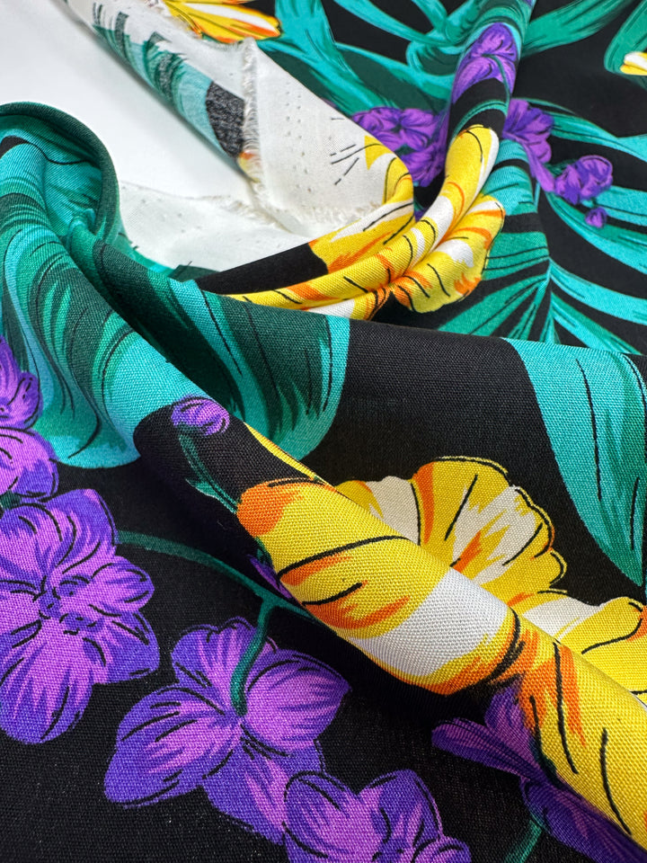 A close-up of Super Cheap Fabrics' Designer Rayon - Neon Canopy (145cm), featuring a lightweight rayon with tropical yellow and white flowers, purple orchids, and teal leaves on black, partially folded to show both front and back.