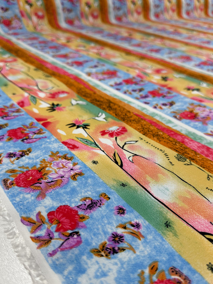 Close-up of Super Cheap Fabrics Designer Rayon - Wildframe (145cm) featuring vibrant floral patterns in red, pink, and purple on a blue, yellow, and orange background with textured rows and fringe edging.