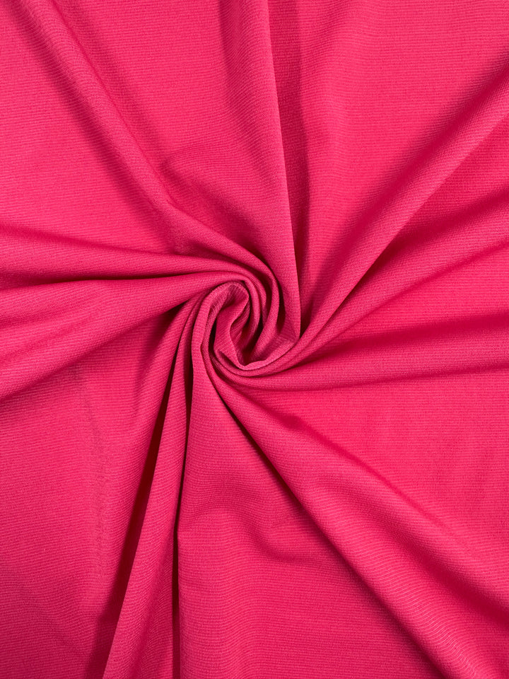 The ITY Knit - Bright Rose - 150cm from Super Cheap Fabrics features a soft, medium weight fabric in bright rose, twisted into a spiral pattern at the center with gentle folds and shadows radiating outward.