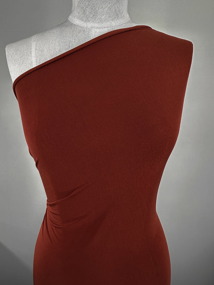 A dress form showcases a fitted, one-shoulder dress made from Super Cheap Fabrics’ ITY Knit - Ketchup (150cm). The rust-colored polyester spandex fabric features smooth lines and subtle waist ruching, set against a plain, light gray background.