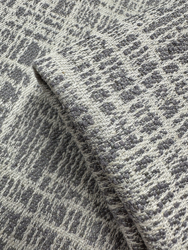 A close-up of Super Cheap Fabrics’ Textured Weave Upholstery - Dexter 33 Dove (147cm), showing its gray and white irregular geometric pattern, folded to display the fabric’s thickness and detailed weave.