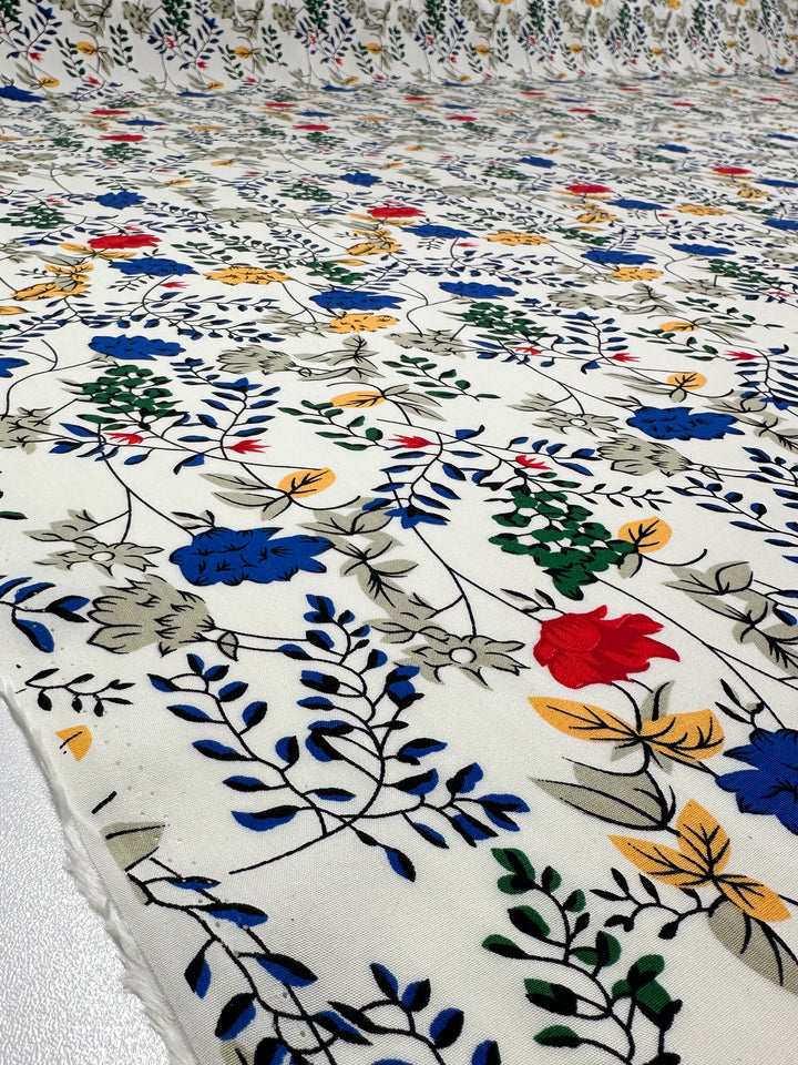 A close-up of Super Cheap Fabrics' Designer Rayon - Petal Grid (145cm), showcasing lightweight rayon with a colorful blue, red, yellow, and green floral pattern and black outlined leaves on a white background.