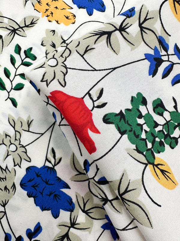 A close-up of Super Cheap Fabrics’ Designer Rayon - Petal Grid (145cm) shows its lightweight rayon with a floral print in red, blue, yellow, green, and black stems on folded fabric, highlighting the colorful botanical design.