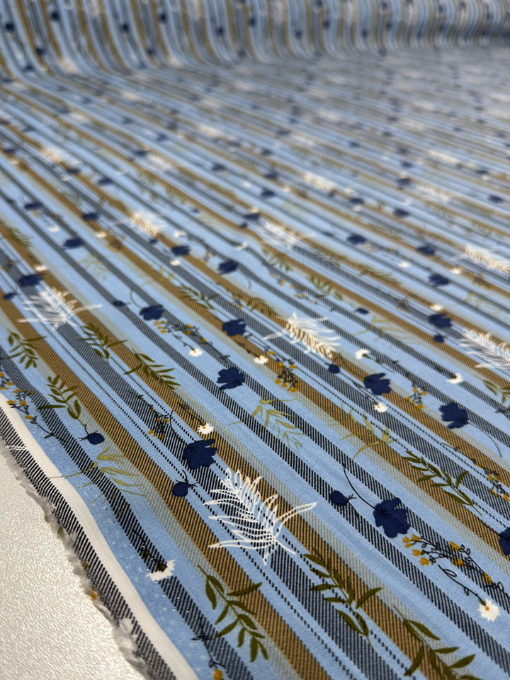 A close-up of Super Cheap Fabrics’ Designer Rayon - Fieldstripe Bloom (145cm), showing colourful rayon with brown, white, and black vertical stripes plus small floral and leaf patterns in white, navy, and green. The fabric edge is visible on the left.