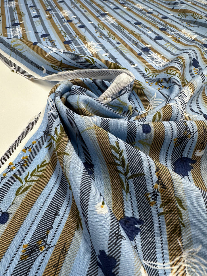 A close-up of Super Cheap Fabrics' Designer Rayon - Fieldstripe Bloom (145cm), showing light blue fabric with diagonal brown, blue, and white stripes, plus a floral and leaf pattern, softly gathered and draped on a flat surface.