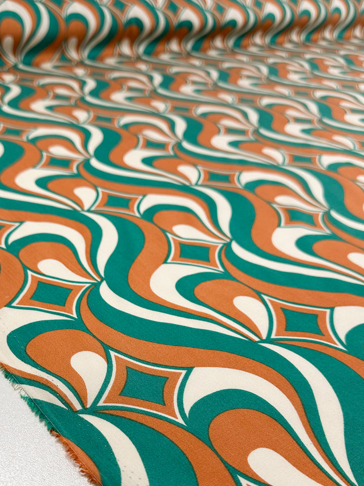 Super Cheap Fabrics’ Designer Rayon - Retro Pulse (145cm) features a close-up of its retro geometric pattern with flowing orange, green, and cream shapes, plus repeating diamond motifs.