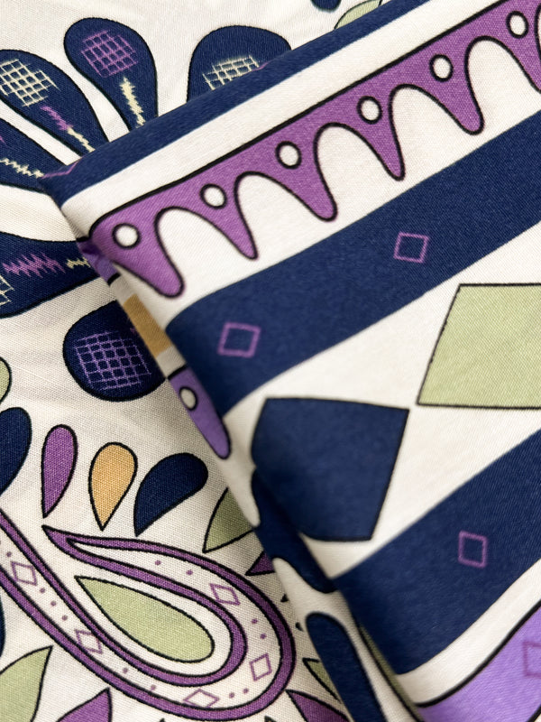 A close-up of Super Cheap Fabrics’ Designer Rayon - Lilac Tempo (145cm), slightly folded, showing vibrant navy blue, purple, green, and yellow geometric and paisley patterns with stripes, diamonds, teardrops, and curves.