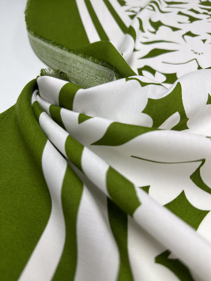 A close-up of Super Cheap Fabrics' Designer Rayon - Olive Fade (145cm) shows smooth folds, visible texture, and bold olive green patterns on a white background, with fabric edges in view.
