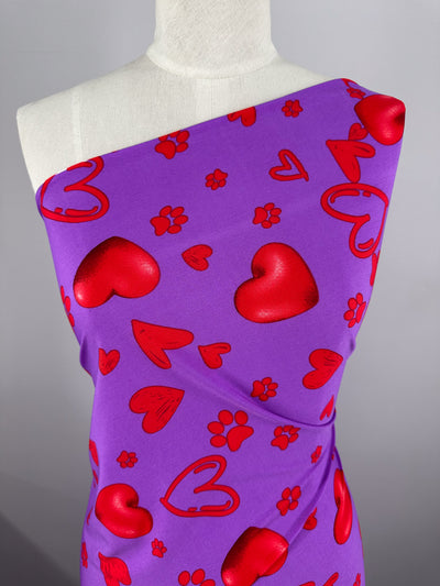 A mannequin displays Super Cheap Fabrics’ Printed Lycra - Love Riot - 150cm, draped over one shoulder to highlight its bold red heart and paw print design on medium-weight lycra for a colorful, playful look.
