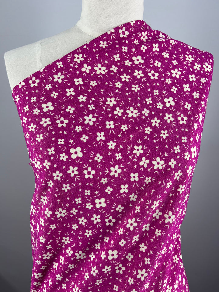 A dressmaker's mannequin draped with a vibrant magenta Bamboo Rayon - Pink DaisyRain - 145cm fabric from Super Cheap Fabrics featuring a pattern of small white flowers scattered across its surface. The background is plain and gray, highlighting the bright, floral and lightweight fabric.