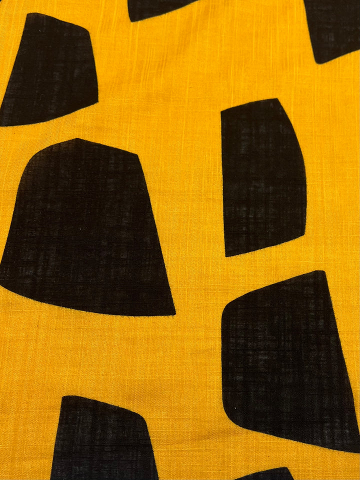 The Bamboo Rayon - Bedrock fabric from Super Cheap Fabrics, measures 152cm and features a yellow lightweight design adorned with irregular black geometric shapes. The shapes resemble trapezoids with curved edges, forming a modern and abstract pattern that's perfect for beginners.