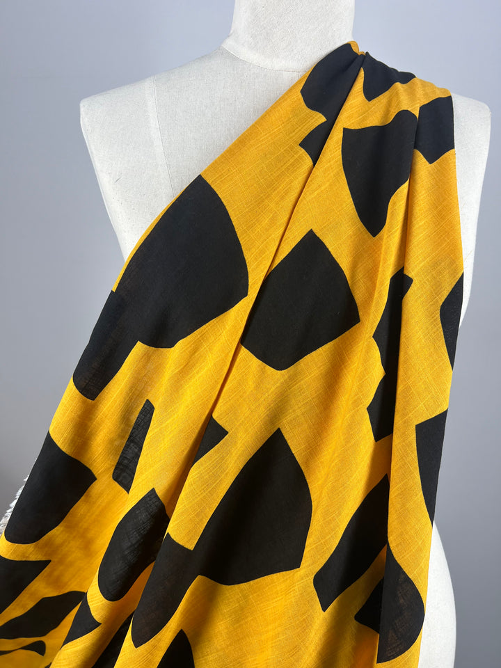 Close-up of a white mannequin draped with Super Cheap Fabrics' Bamboo Rayon - Bedrock - 152cm, a yellow, lightweight fabric featuring a bold black abstract pattern. The vibrant Bamboo Rayon fabric showcases large irregular shapes that create a striking contrast against the yellow background. The backdrop is a simple gray gradient.