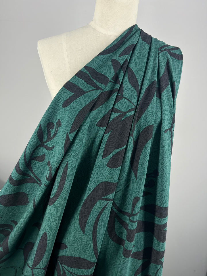 A mannequin draped in the "Bamboo Rayon - Landscape - 152cm" from Super Cheap Fabrics showcases the teal bamboo rayon featuring an abstract black leaf pattern. This lightweight fabric is gathered gracefully over one shoulder, cascading down in elegant folds. The plain white background enhances the design and texture, making it an ideal choice for home decor enthusiasts.