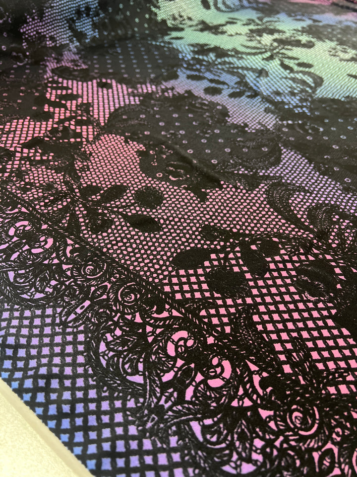 Close-up of the Printed Lycra - Laced by Super Cheap Fabrics, showcasing a medium-weight Polyester/Spandex blend with an intricate black lace pattern overlaying a multicolored iridescent background. The design features floral and geometric elements that create a striking contrast between the delicate black lace and the vibrant hues underneath. This fabric is available at a width of 150cm.