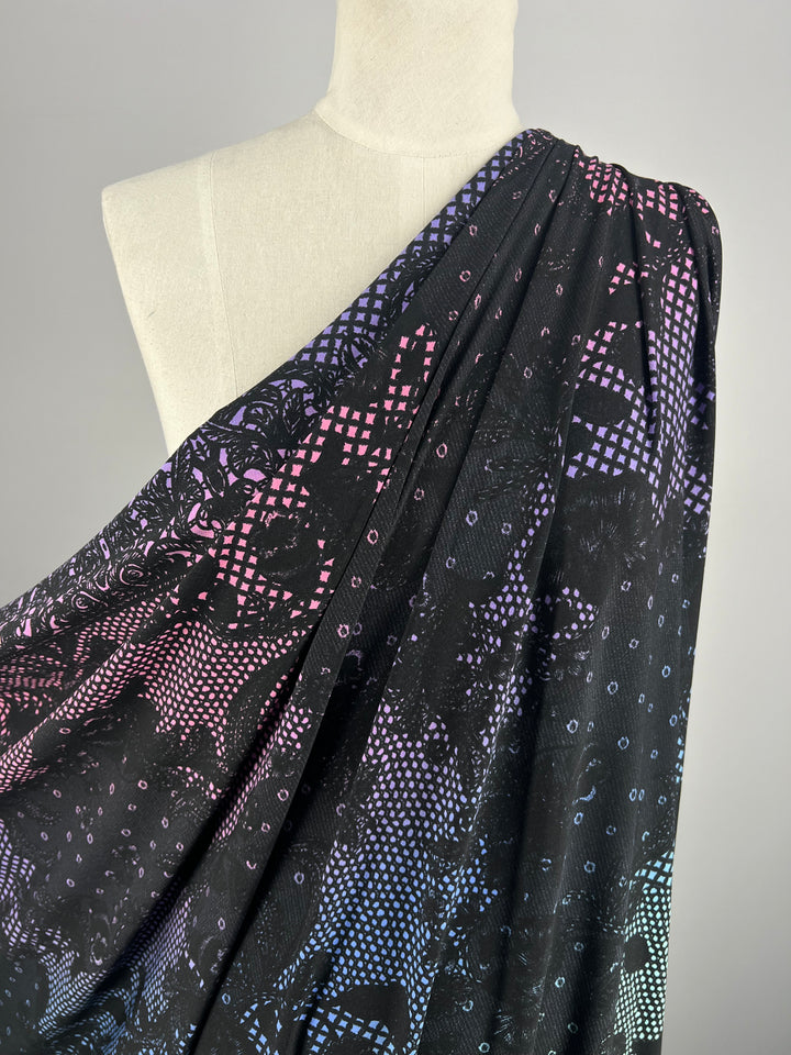 A mannequin draped in Super Cheap Fabrics' "Printed Lycra - Laced - 150cm," a black, medium-weight Polyester/Spandex fabric adorned with a geometric pattern featuring small squares in shades of pink, purple, and blue that create a gradient effect across the material. The fabric hangs over one shoulder, showcasing its texture and design.