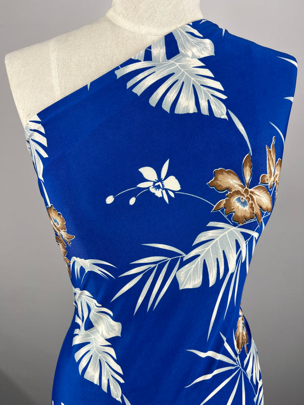 A mannequin is draped in Super Cheap Fabrics' vibrant blue medium-weight Printed Lycra - Tropski, featuring a white and brown tropical floral pattern. The floral design includes large leaves and blossoms, creating a striking contrast against the blue background. The fabric is arranged in a one-shoulder style.