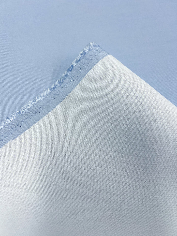 Close-up image of Suiting - Clear Sky - 142cm by Super Cheap Fabrics, featuring a light blue fabric with a folded corner that reveals its selvage edge. The medium weight material appears smooth and slightly shiny, indicating either a high thread count or a synthetic blend.