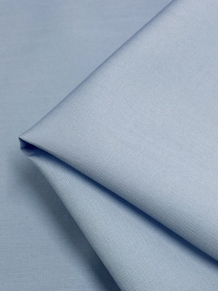 A close-up photo of the medium weight "Suiting - Clear Sky - 142cm" fabric by Super Cheap Fabrics, showcasing a light blue, smooth texture. Folded neatly with distinct creases and shadows, this 97% cotton fabric appears soft and pristine, highlighting its fine weave and uniform color.