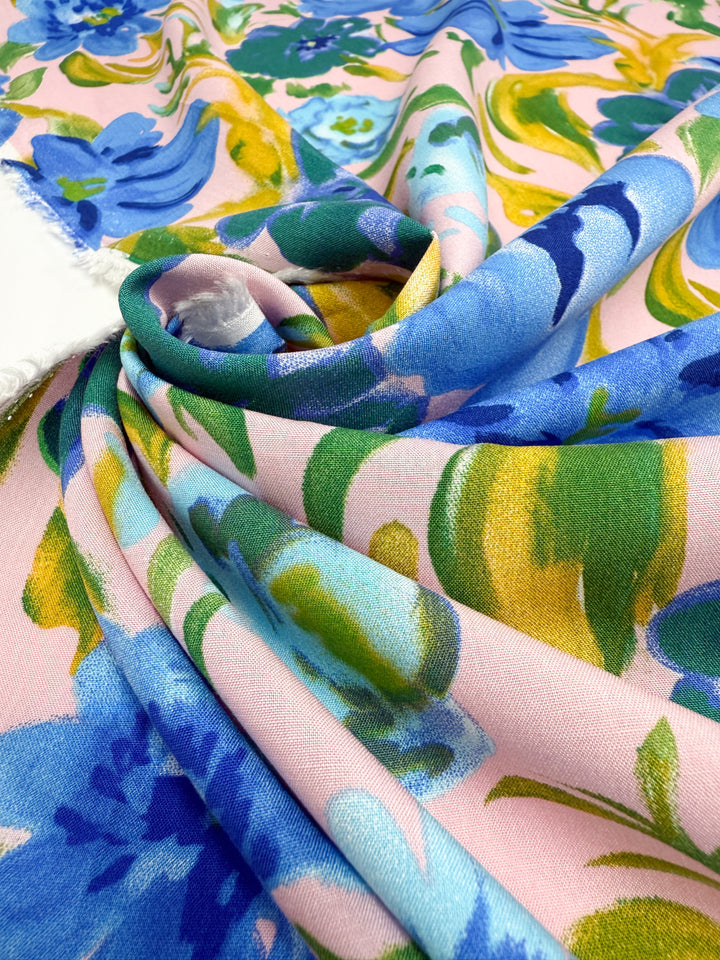 A close-up of Super Cheap Fabrics' Designer Rayon - Bluebell Blush (145cm), featuring vibrant blue, green, and yellow floral patterns on light pink, with an organic drape and visible white selvage edge.