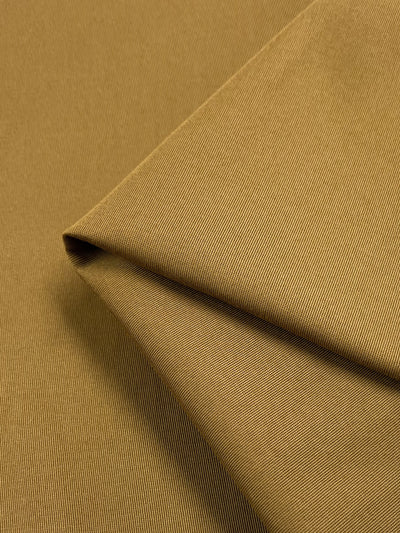 A close-up photo of Gabardine Suiting in Cumin from Super Cheap Fabrics reveals a golden-brown, medium to heavy weight cotton fabric with a fine, diagonal ribbed texture. The 140cm wide fabric is folded over itself, creating clean, sharp lines and showcasing the material's smooth surface and rich cumin color, ideal for stylish pants.