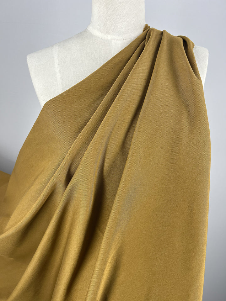 The Gabardine Suiting fabric in a warm cumin shade from Super Cheap Fabrics is draped elegantly around a white dress form. This medium to heavy weight cotton material features soft folds that create a sense of fluidity and movement. The plain and neutral background highlights the texture and rich color of the 140cm fabric, making it ideal for stylish pants.