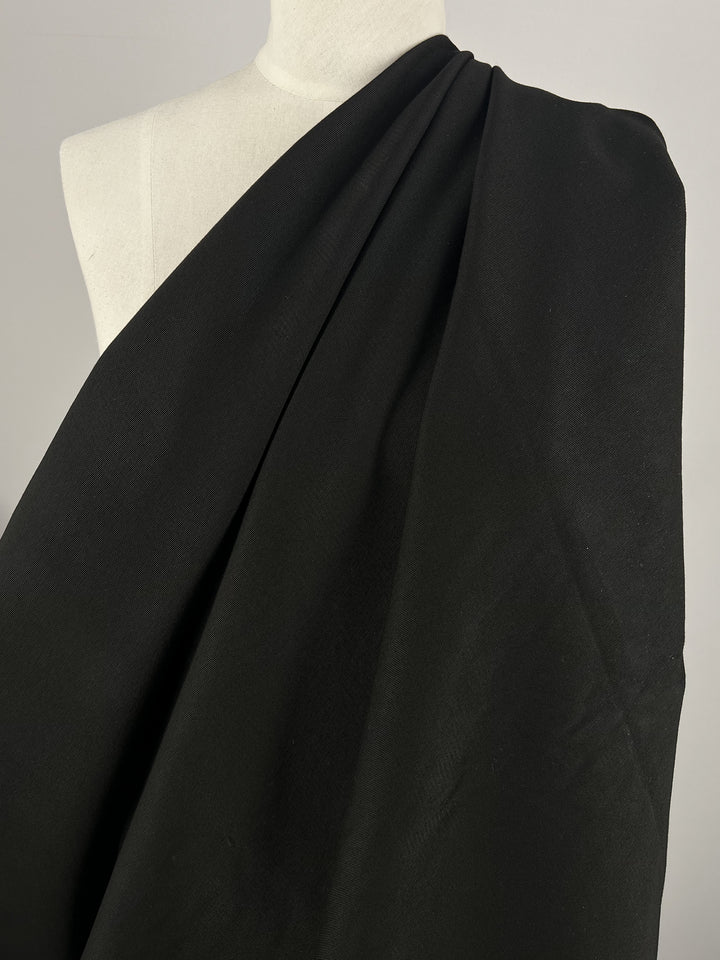 A close-up of a mannequin draped in Super Cheap Fabrics' Gabardine Suiting in black, 140cm wide. The medium to heavy weight cotton material is arranged to cover the left shoulder and drapes diagonally across the chest, leaving the right shoulder exposed. The plain, light-colored background highlights the dark fabric beautifully.