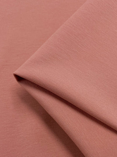 A close-up view of a folded piece of soft, textured fabric in a muted peach or dusty pink color, referred to as Coral Cloud. The Milano Ponte fabric from Super Cheap Fabrics appears smooth and lightweight, with subtle weave patterns visible. The overlapping layers create gentle shadows and a sense of depth. This Medium Weight Ponte Fabric measures 138cm wide and offers an elegant drape.