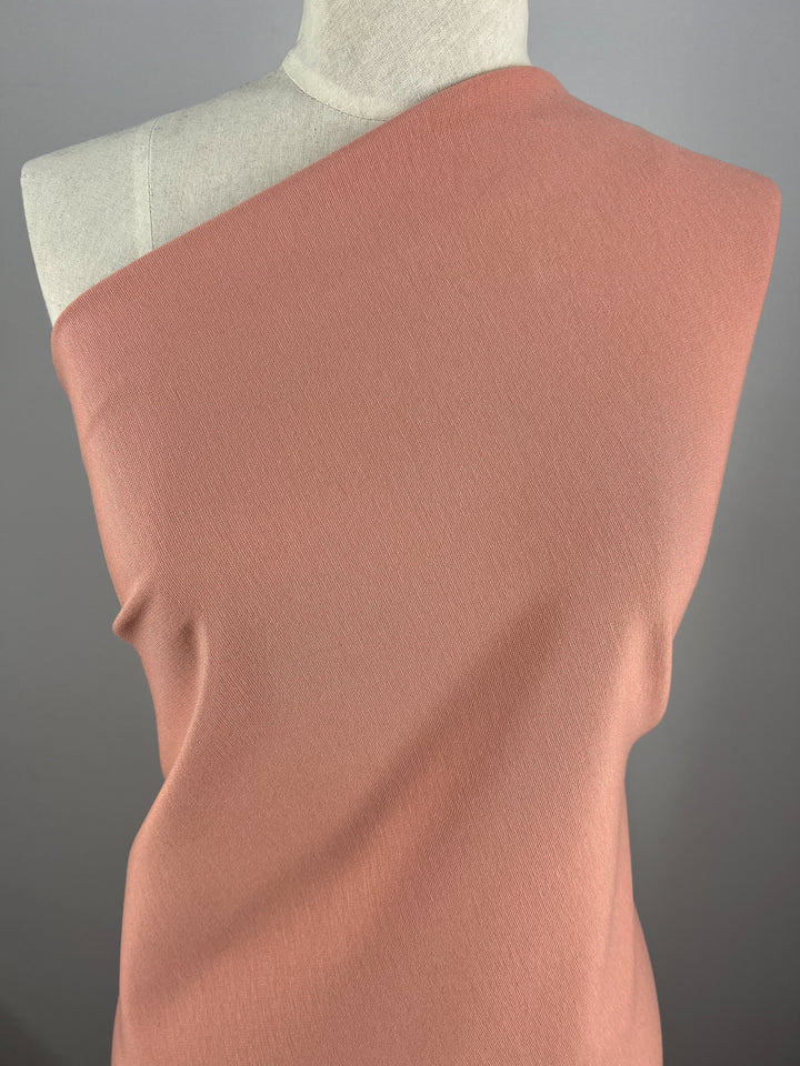 A close-up view of a mannequin wearing a one-shoulder, sleeveless top made from Super Cheap Fabrics' Milano Ponte in Coral Cloud - 138cm. The background is a plain, neutral grey, which accentuates the garment's texture and vibrant color.