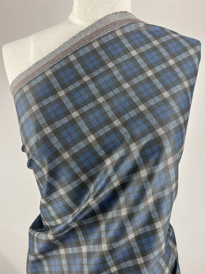 A mannequin is draped with a piece of Super Cheap Fabrics' Ponte - Checkers fabric, measuring 150cm and exhibiting a medium-weight structure in grey, blue, and black plaid. The checkered pattern features alternating blue and black squares intersected by thin grey lines, making it an ideal choice for pants. The edges of this polyester fabric are adorned with a red and white stitch pattern.