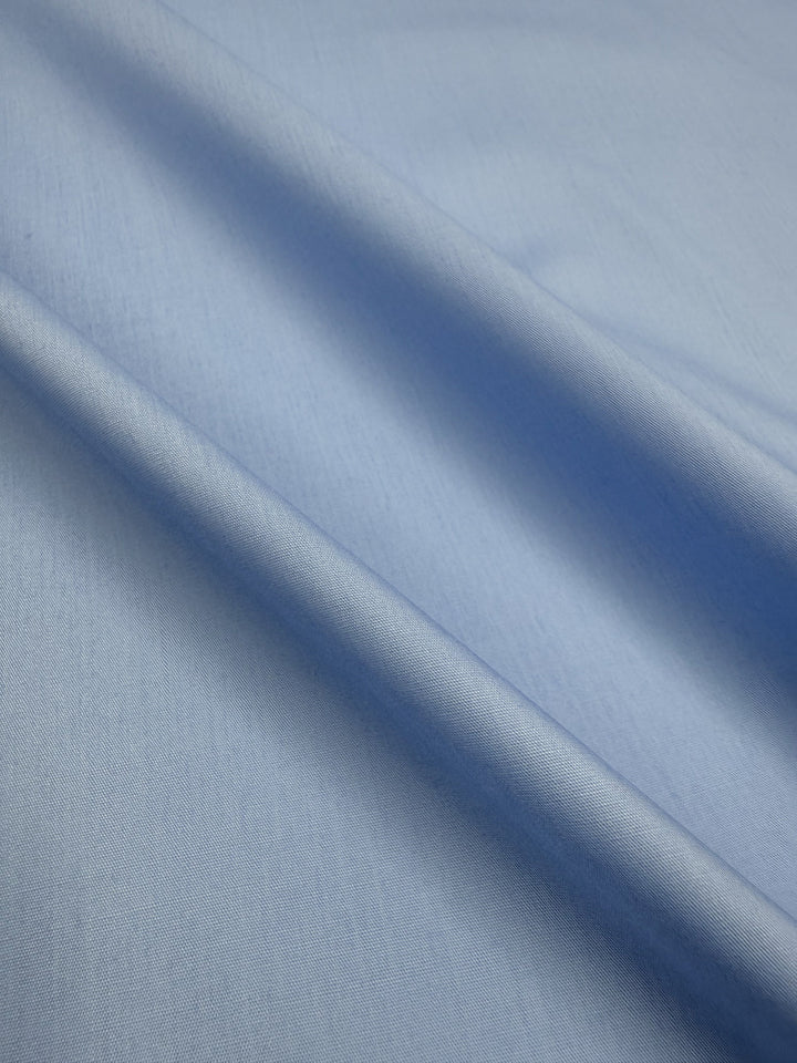 A close-up of Super Cheap Fabrics' Poplin - Powder Blue - 114cm, showcasing its smooth, lightweight poly cotton fabric with soft diagonal folds that create gentle shadows and enhance its texture.
