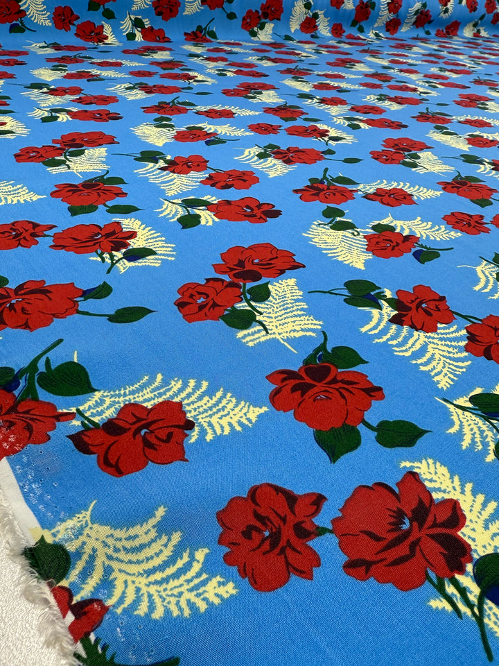 Designer Rayon - Rose Rapture by Super Cheap Fabrics is a blue rayon fabric (146cm wide) featuring vibrant red roses, green leaves, and pale yellow fern-like patterns, displayed flat on a surface.