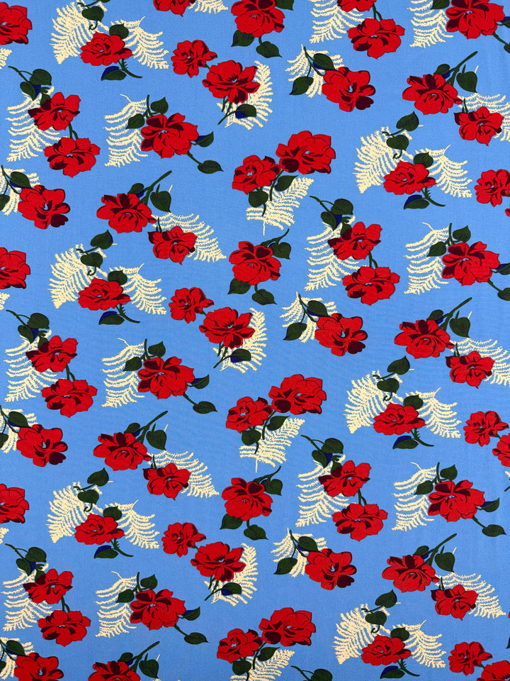 Super Cheap Fabrics' Designer Rayon - Rose Rapture (146cm) features a repeating pattern of red roses and cream fern-like leaves scattered over a bright blue background.