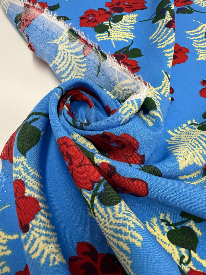 Designer Rayon - Rose Rapture by Super Cheap Fabrics (146cm) features a blue base with red flowers and cream ferns, shown flat and partly rolled to highlight its vibrant design and smooth texture.