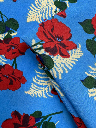 A close-up of Super Cheap Fabrics’ Designer Rayon - Rose Rapture (146cm) shows bold red flowers, green leaves, and cream ferns on soft rayon, with vibrant colours highlighted by gentle folds in the fabric.