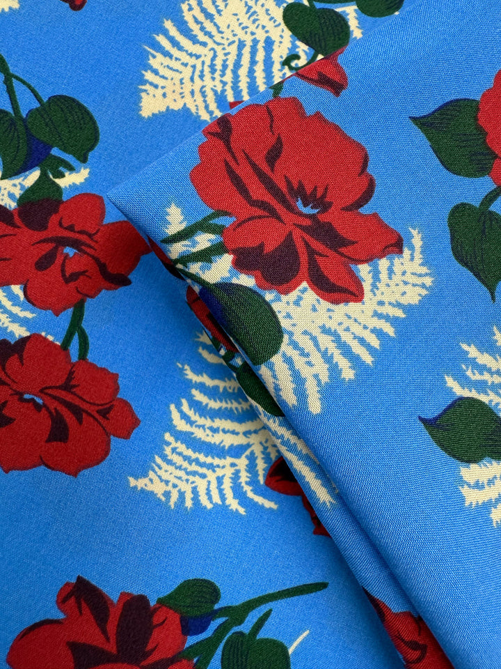 A close-up of Super Cheap Fabrics’ Designer Rayon - Rose Rapture (146cm) shows bold red flowers, green leaves, and cream ferns on soft rayon, with vibrant colours highlighted by gentle folds in the fabric.
