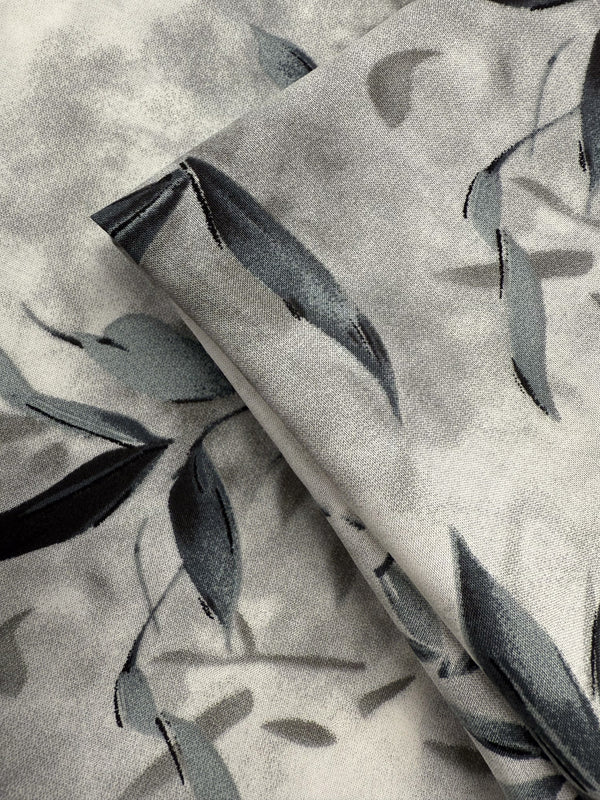 Close-up of Super Cheap Fabrics' Designer Rayon - Misted Olive (147cm), showcasing a soft watercolor-style print with gray and black leaves and branches on a light gray background.
