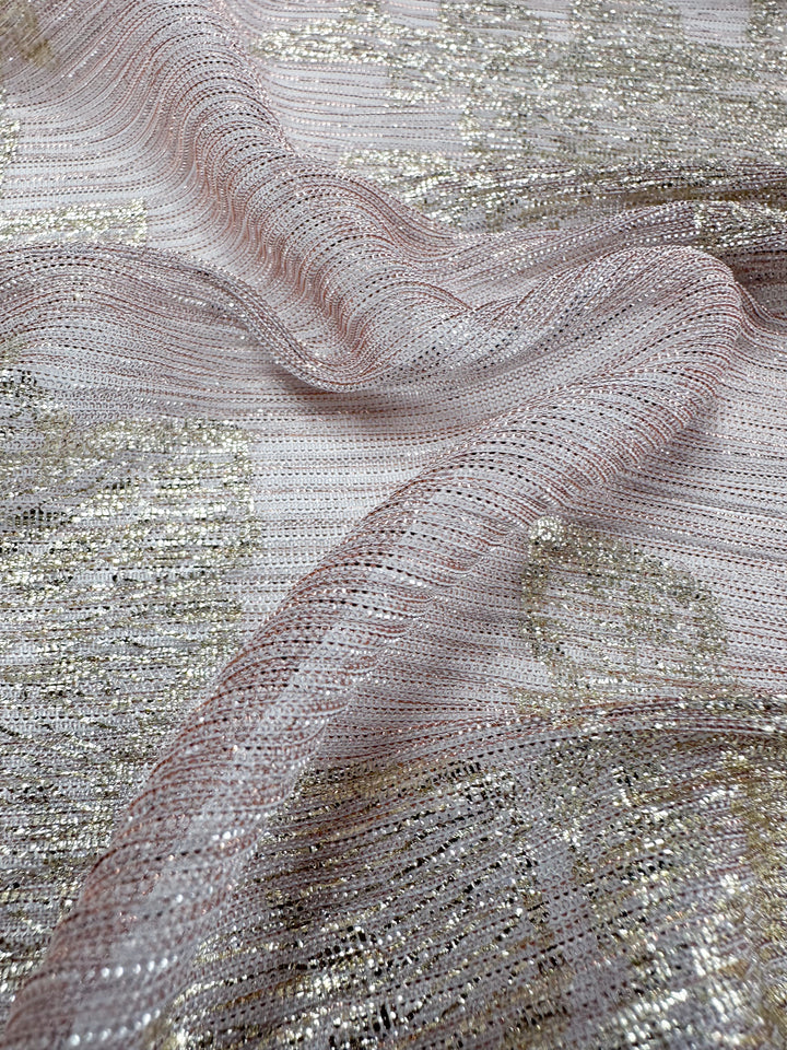 A close-up of the Metallix Pleat - Gold Leaf - 140cm polyester fabric from Super Cheap Fabrics reveals its light pink base, intricately interwoven with metallic silver and gold threads. The lightweight material has a soft, shimmery sheen with gentle folds that enhance its delicate appearance.