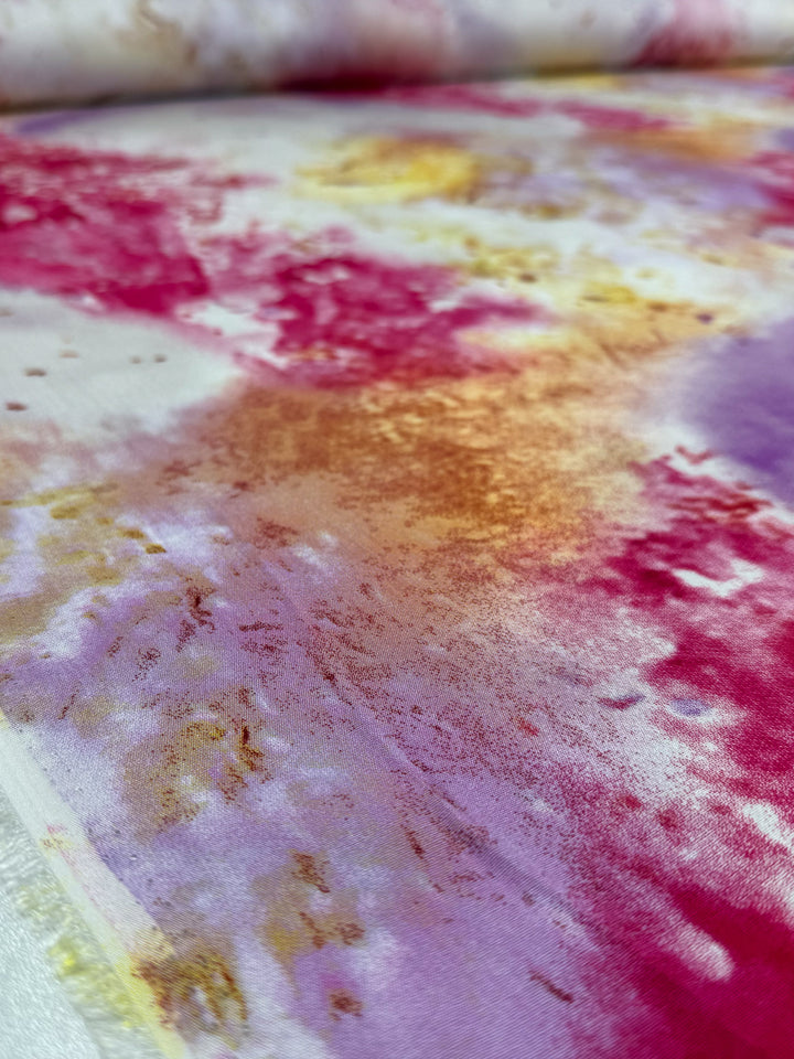 A close-up of Super Cheap Fabrics' Designer Rayon - Aurora Bloom (143cm), showing its vibrant watercolor-style red, yellow, gold, orange, and purple pattern blending on a white background with visible fabric texture.